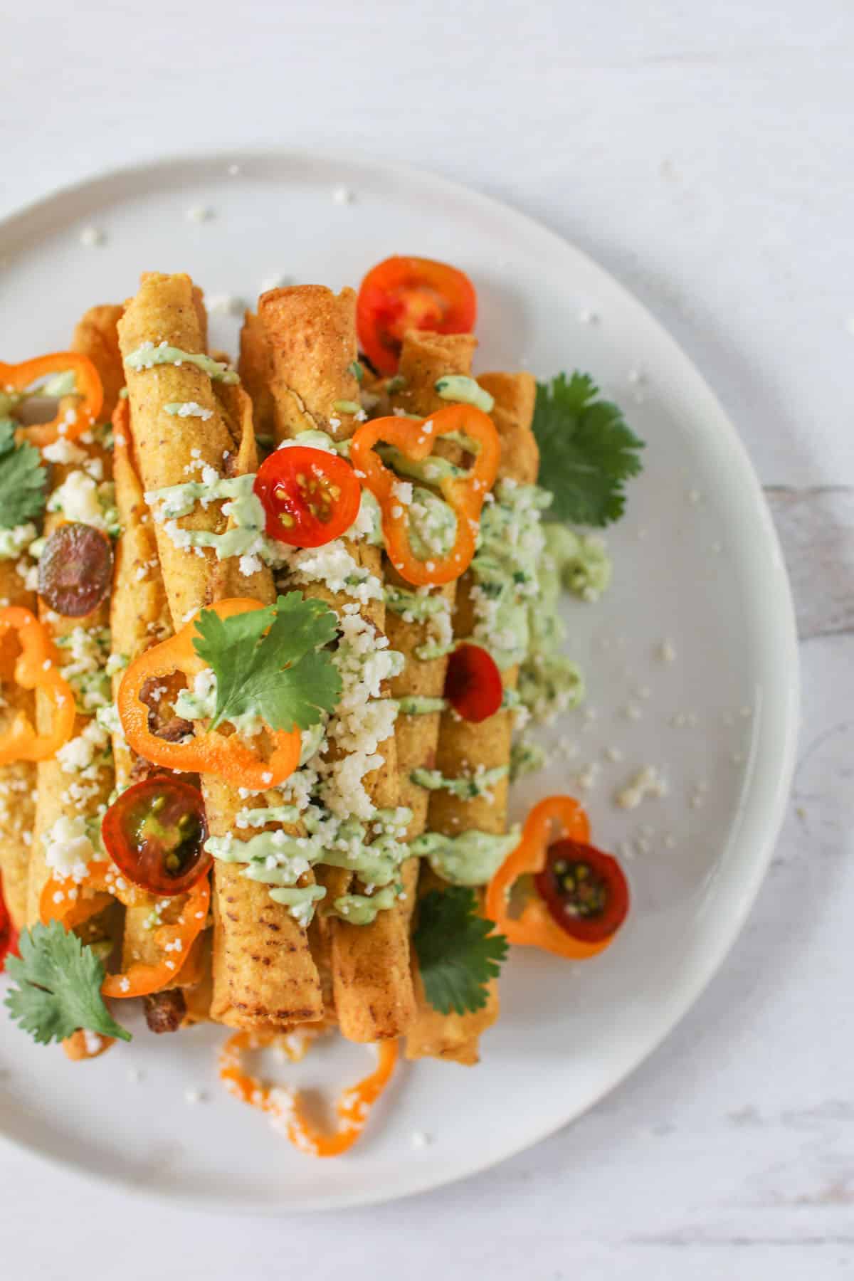 An overhead of crispy taquitos stacked on a white plated drizzled with avocado cream sauce and topped with cilantro, cojita cheese, sliced cherry tomatoes and peppers.