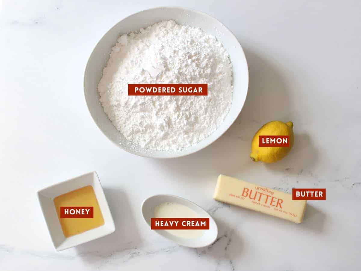 Lemon Honey Frosting ingredients in white bowls except for 1 whole lemon and 1 stick of butter in the originally wrapping. Each ingredient is labeled in a red box with white text.