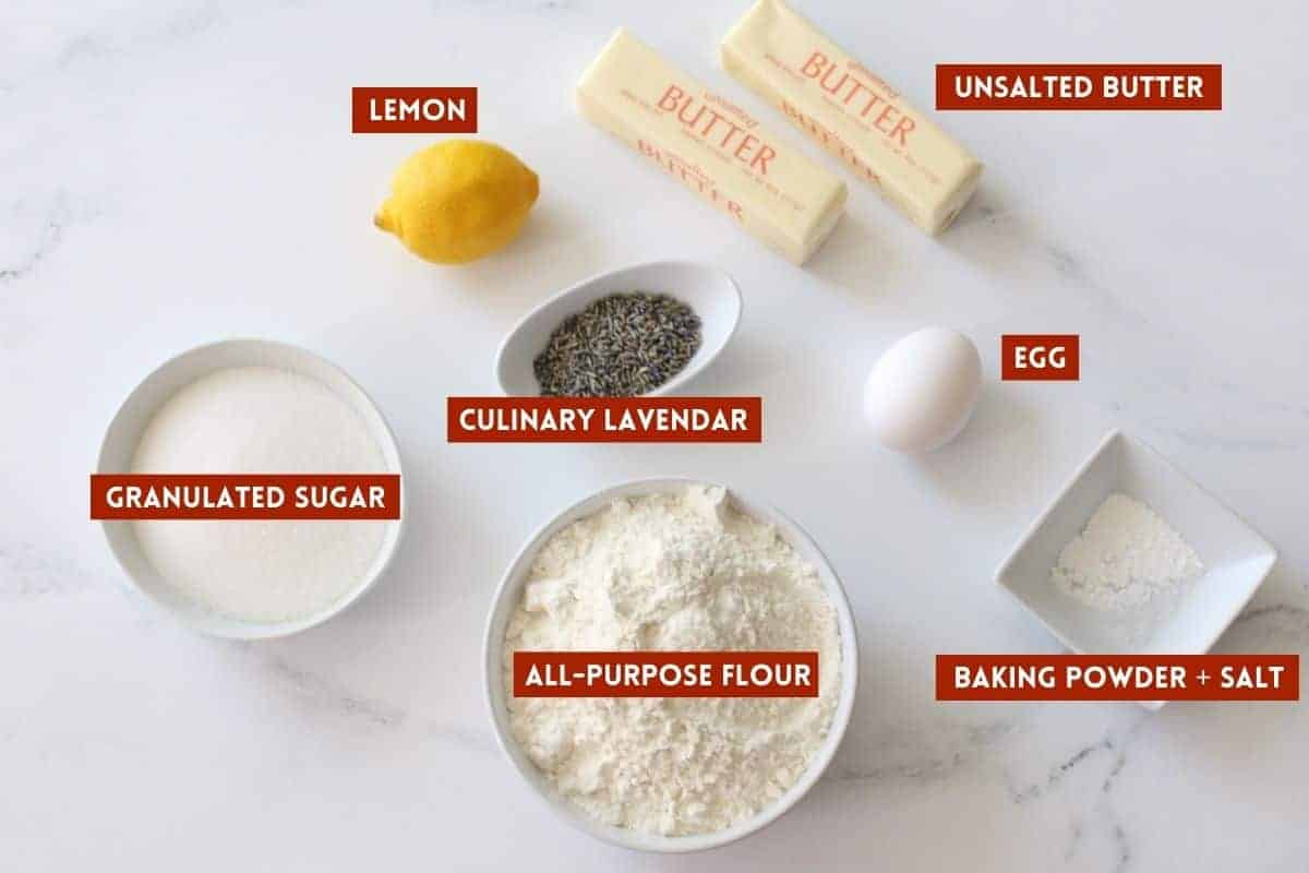 Lemon Lavender Cookie Bar Ingredients in different sized white bowls except for 1 whole lemon, 1 white egg, and 2 sticks of butter in the wrapping paper on a white marble background. Each ingredient is labeled with a red box with white text.