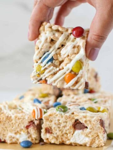A hand holding a Rice Krispies Treat with more M&M Rice Krispie Treats underneath on a white wood board with brown parchment paper.
