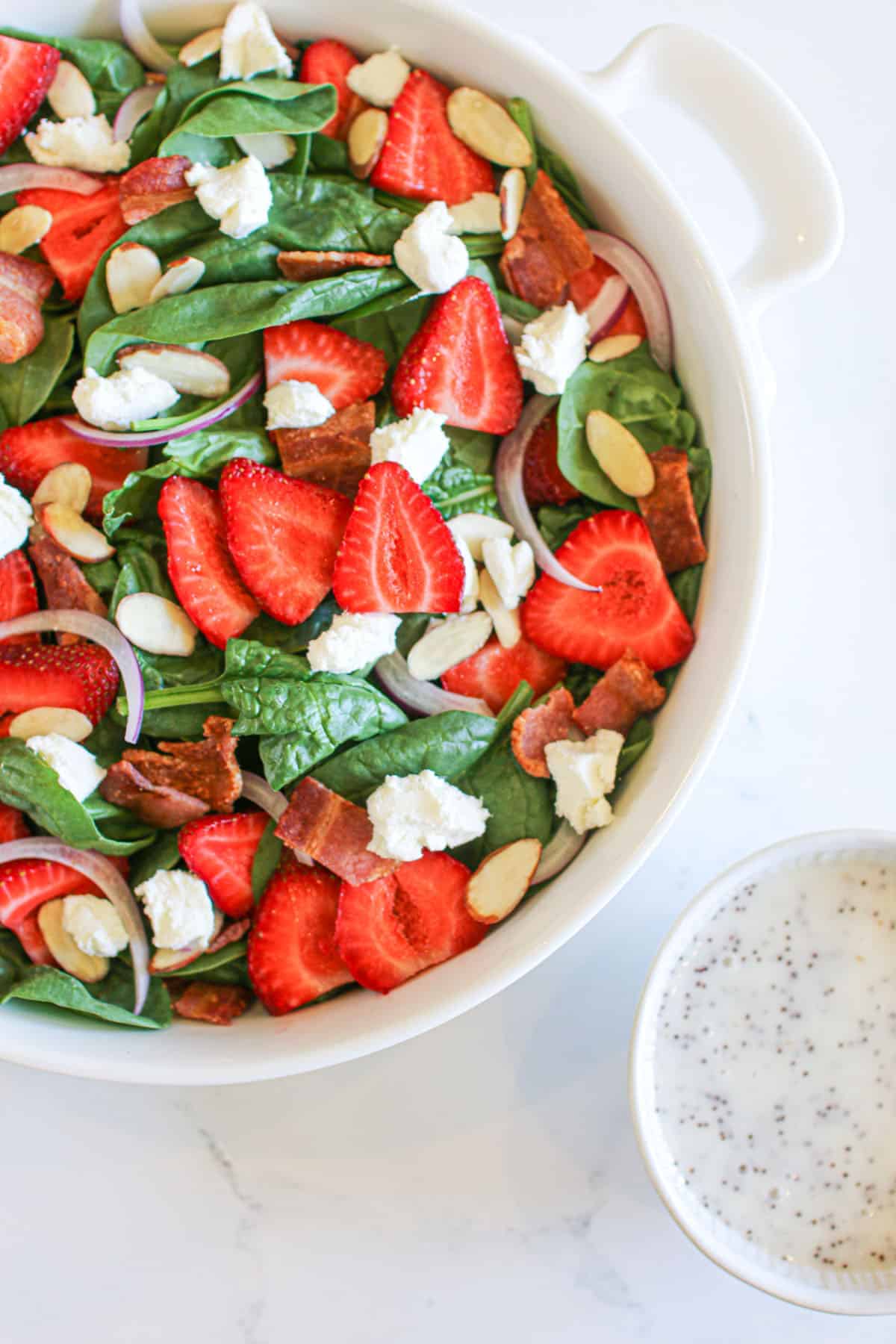 A white bowl with strawberry goat cheese spinach salad on a white marble background with a small bowl off to the side with creamy poppyseed dressing.