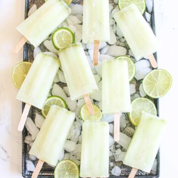 Lime popsicles on a baking sheet with ice cubes and sliced limes on a white marble background.
