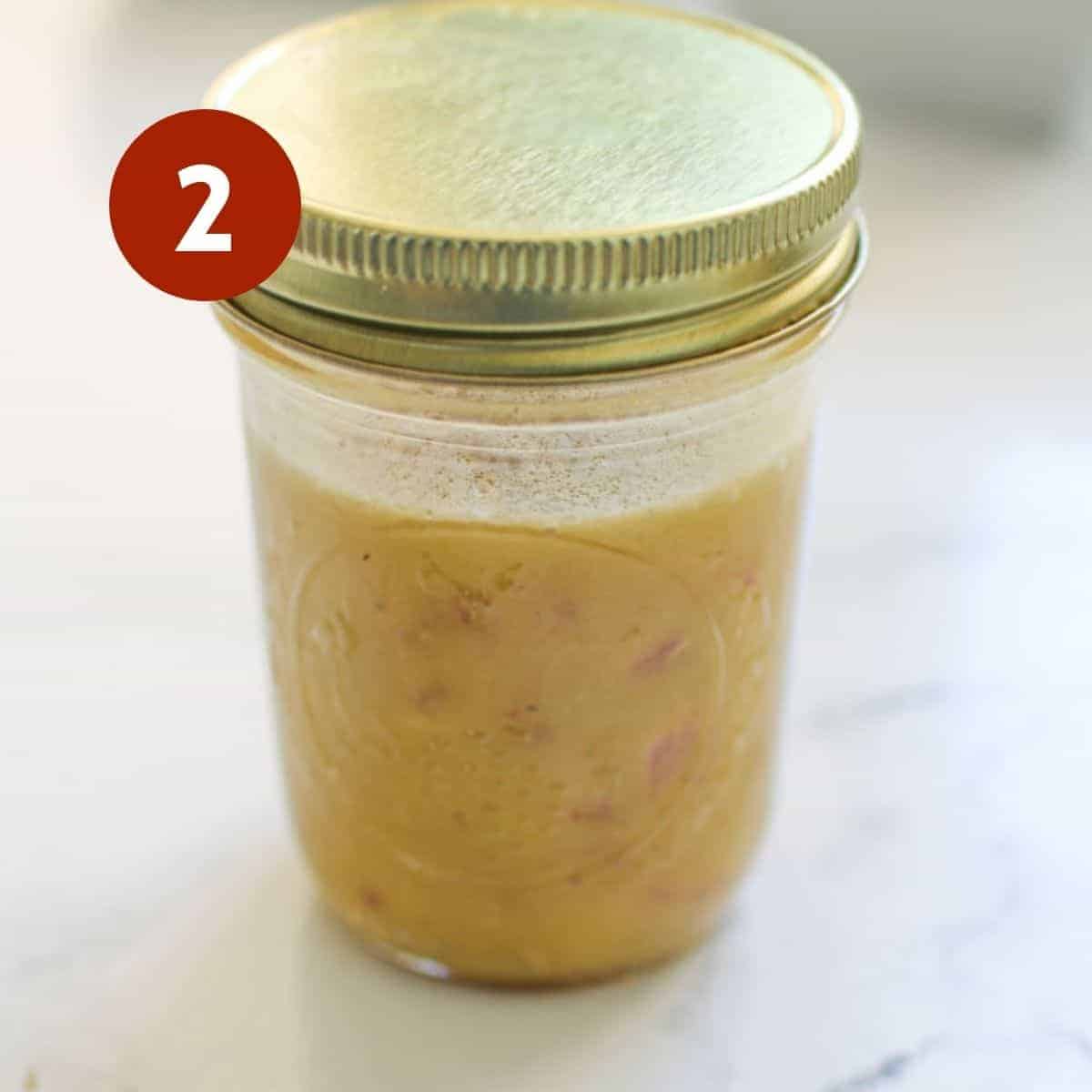 A mason jar with a yellow dressing in it with a gold lid on top on a white marble background. There's a white #2 in a red circle in the upper left corner.