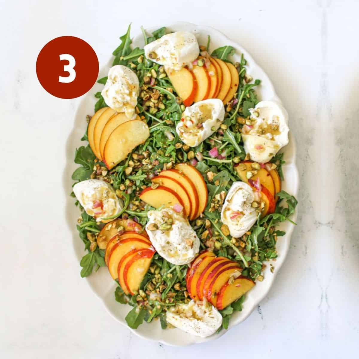 A bed of baby arugula on a white large scallop platter with sliced peaches, torn burrata and crushed pistachios on white marble background. A white #3 in the a red circle in the upper left corner.