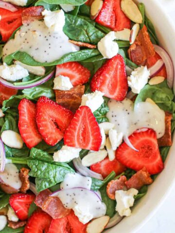 Large white bowl with Spinach Strawberry goat Cheese Salad with sliced almonds, thinly sliced red onion and drops of poppy seed dressing.