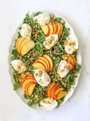 A bed of baby arugula on a white large scallop platter with sliced peaches, torn burrata and crushed pistachios on white marble background.