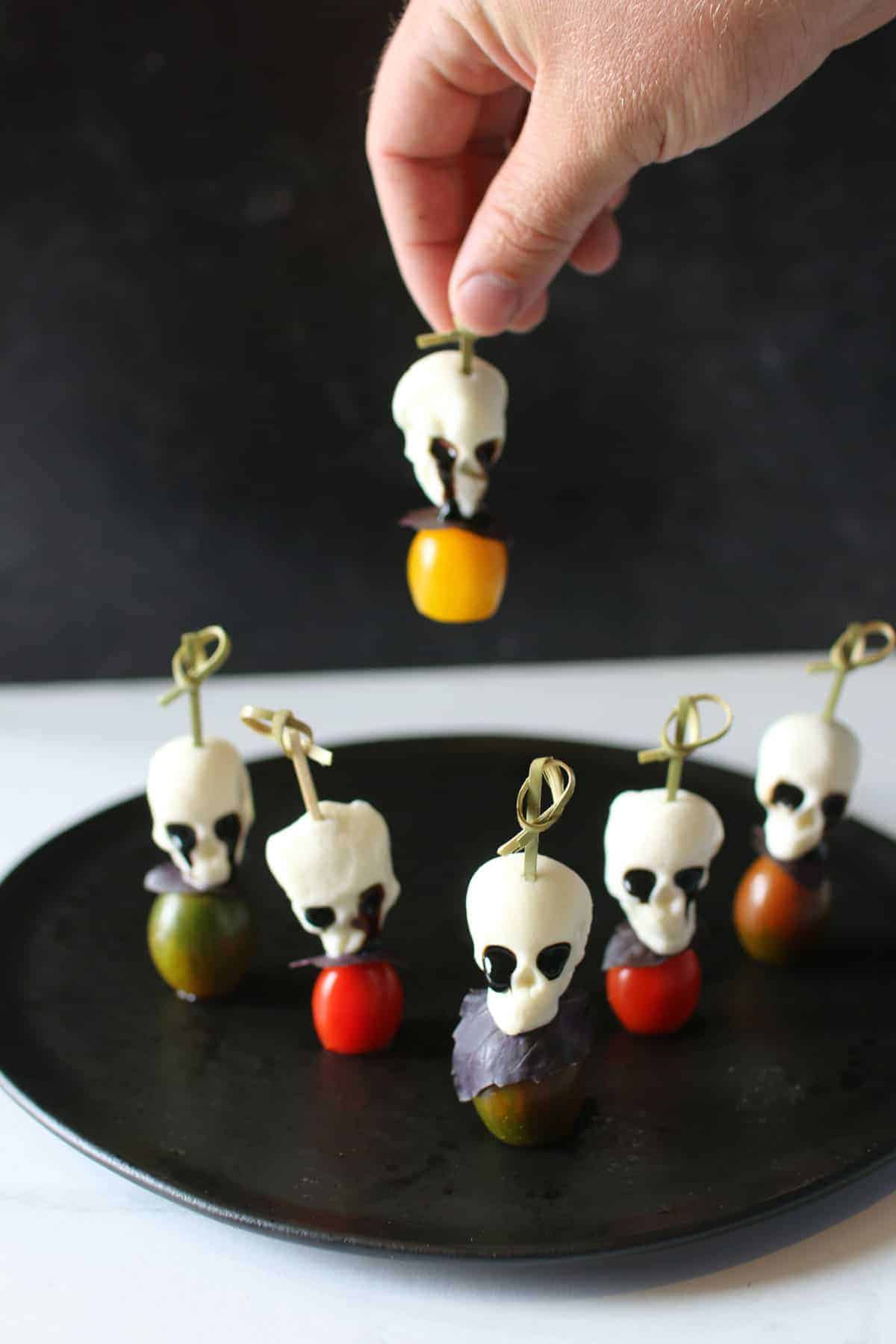 6 Halloween Caprese Skewers sit on a black plate with a hand picking up a skewer in the background. Each skewer consists of a bamboo skewer with a knot at the top, then a piece of skull shaped mozzarella with goo-ing black eyes of balsamic glaze, a piece of purple basil and then a colorful cherry tomato.