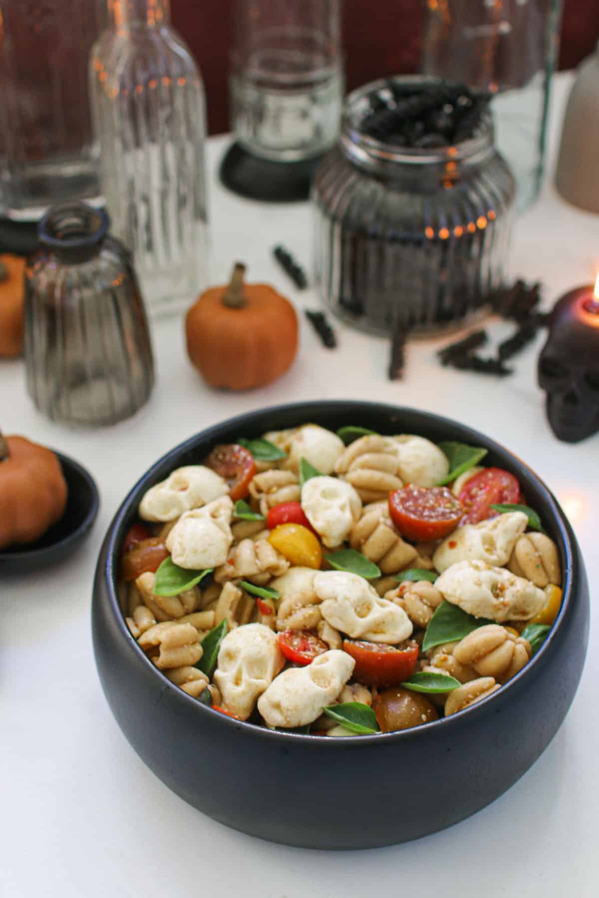 A close up angled photo of Halloween Pasta Salad in a black bowl with pumpkin shaped pasta, mini basil leaves, sliced cherry tomatoes and mozzarella skulls sitting on a white marble table. In the background are assorted decorations of mini orange pumpkins, clear glass bottles and candles with a dark red background.