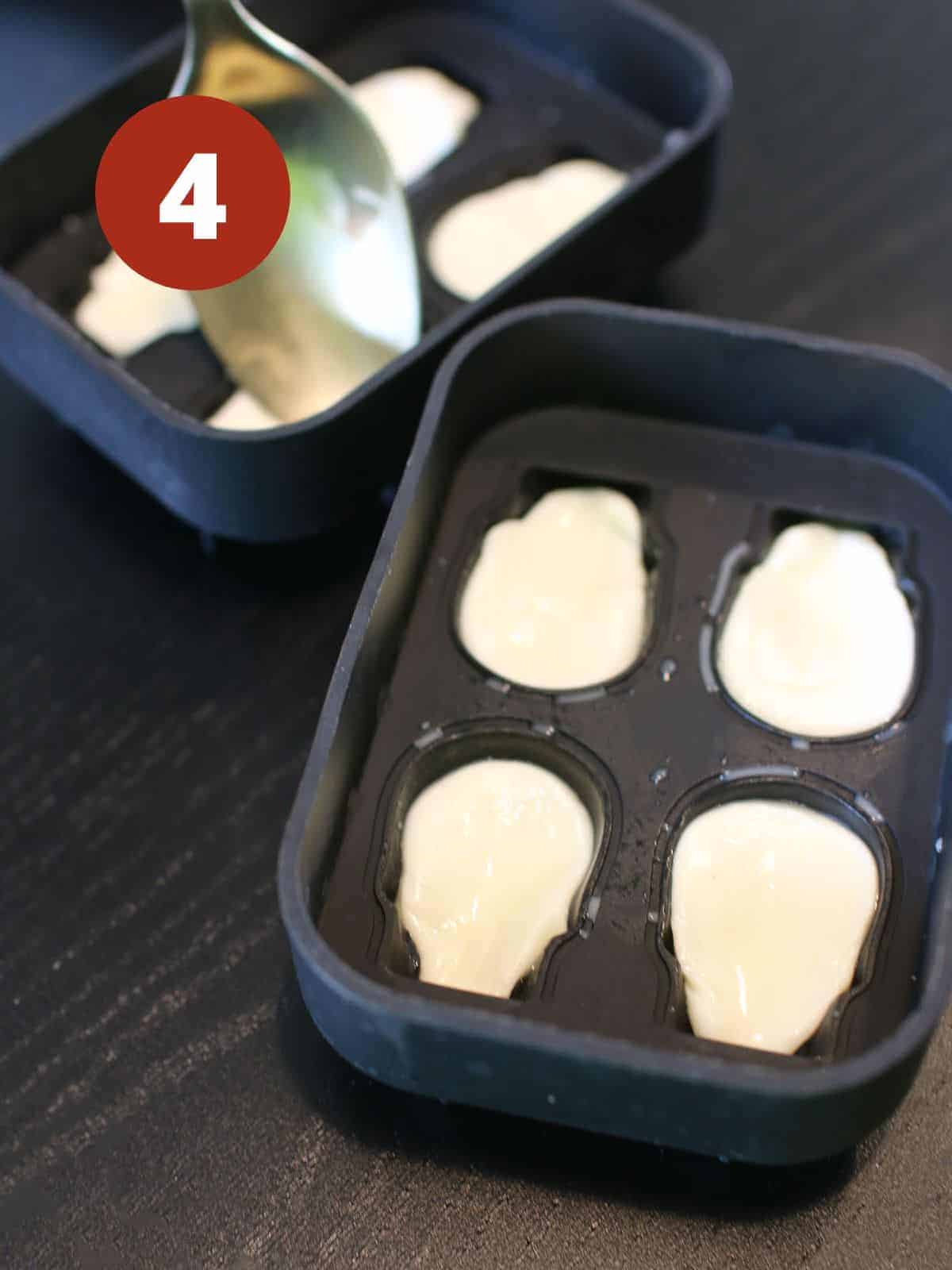 Two skull silicone molds and a mozzarella ball in each crevice. A back of a gold spoon presses mozzarella in the mold crevice. A dark red circle is in the upper left corner and a '4' in white text color.