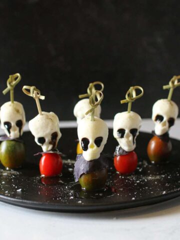6 Halloween Caprese Skewers sit on a black plate with olive oil and flay sea salt on a white marble background with eyes oozing black balsamic glaze
