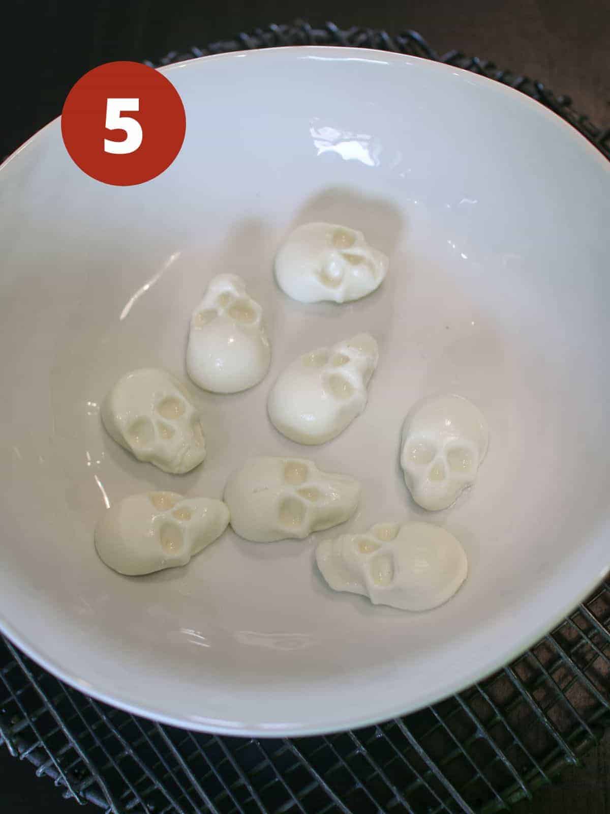 8 mozzarella shaped skulls in a white low bowl on a black wire trivet on a black background. A dark red circle is in the upper left corner and a '5' in white text color.
