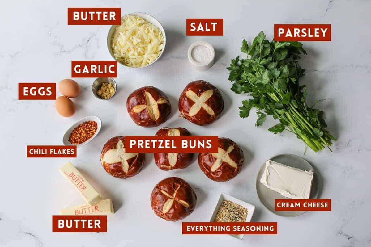 Korean Inspired Everything Cheesy Garlic Pretzel Bread ingredients on a white marble background. Each ingredient is labeled with a dark red rectangle box with white text in all caps.