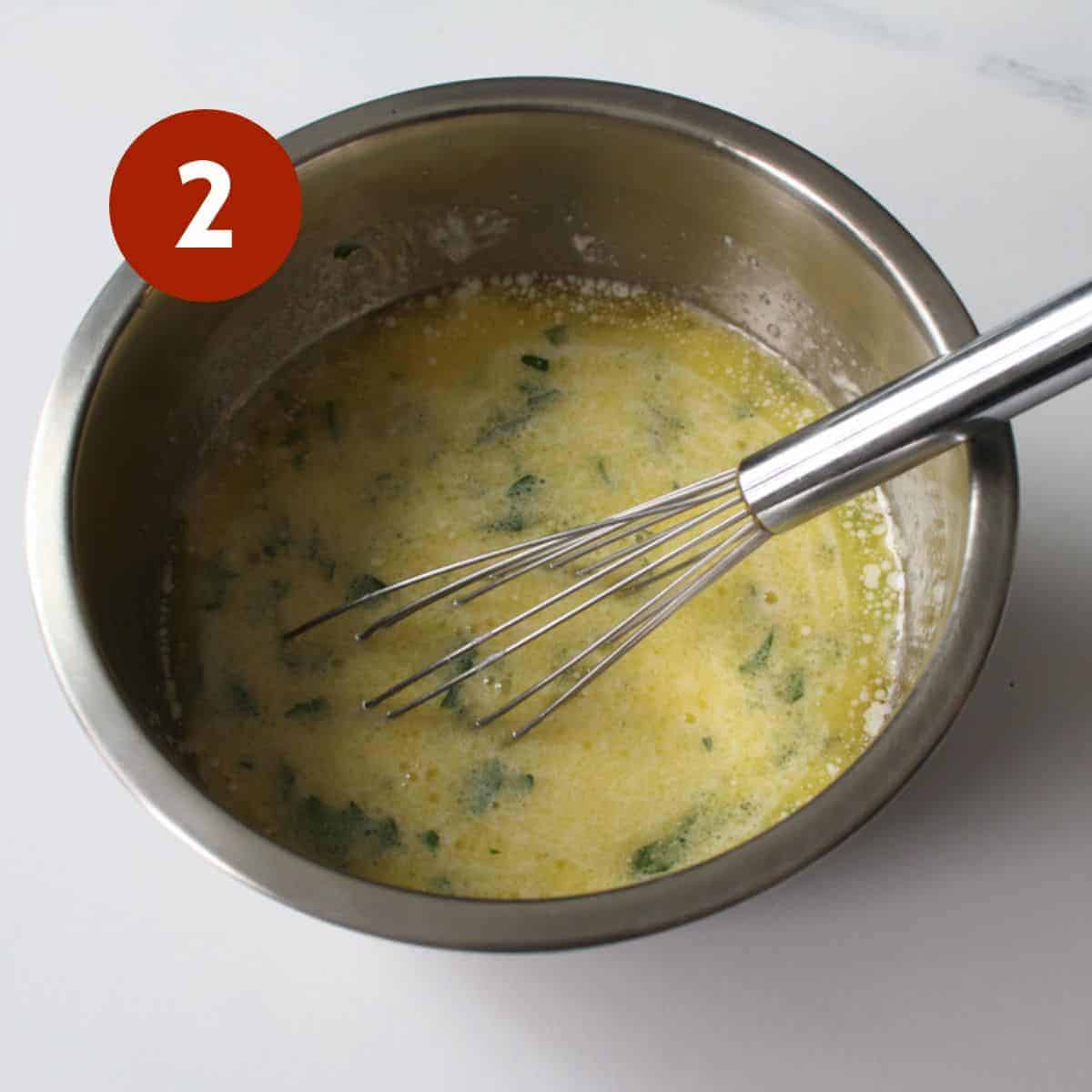 A metal bowl full of garlic butter custard with a metal whisk in the bowl. A dark circle with a white '2' in the upper left corner on a white marble background.