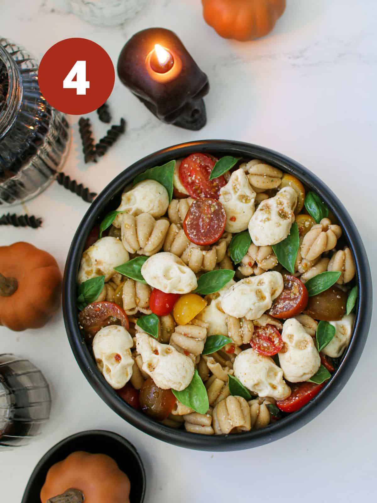 A black bowl of halloween pasta salad on a white background surrounded by mini orange pumpkins, candles, and bottles. In the pasta salad are mini pumpkin shaped pasta, mini basil leaves, sliced cherry tomatoes and skull shaped mozzarella. A white '4' in a dark red circle is in the upper left corner.