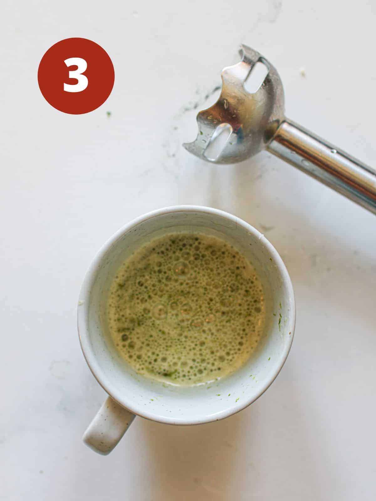 A cup of dressing in a white mug with a hand blender off to the side on a white marble background. A dark red circle with a white '3' is in the upper left corner.