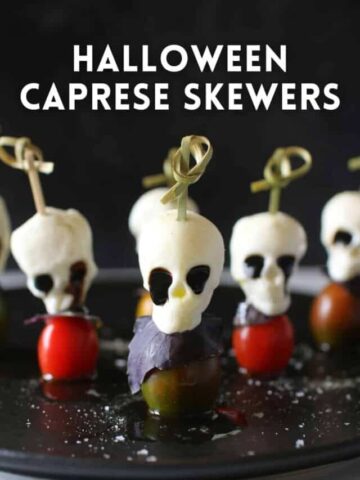 6 Halloween Caprese Skewers sit on a black plate with olive oil and flay sea salt on a white marble background with eyes oozing black balsamic glaze with 'Halloween Caprese Skewers' in white text in all caps in the upper section of the pin and centered.