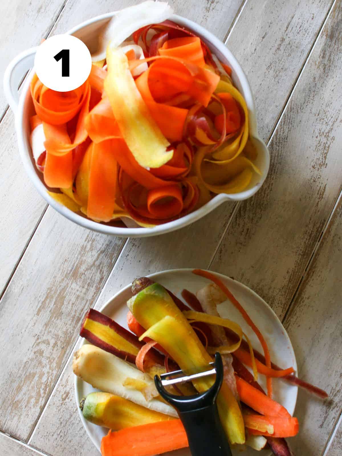 A mixing bowl with colorful carrot ribbons. On a medium plate is a vegetable peeler and carrots being partially peeled on a light brown wood background. A white circle with a black '1' in the upper left corner.