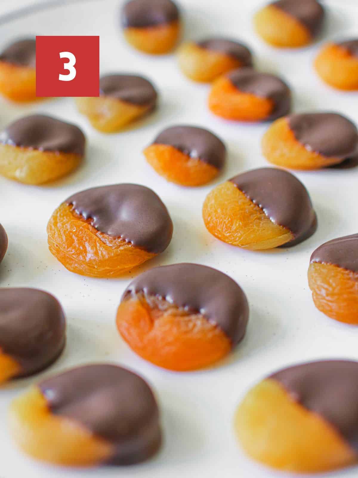 Close up of half dipped chocolate dipped apricots on a white speckled plate. In the upper left corner is a dark red square with a white '2' in the center.