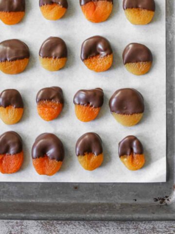 Chocolate Dipped Apricots of all different sizes and half dipped in chocolate on white parchment paper on a sheet pan on a wood background.