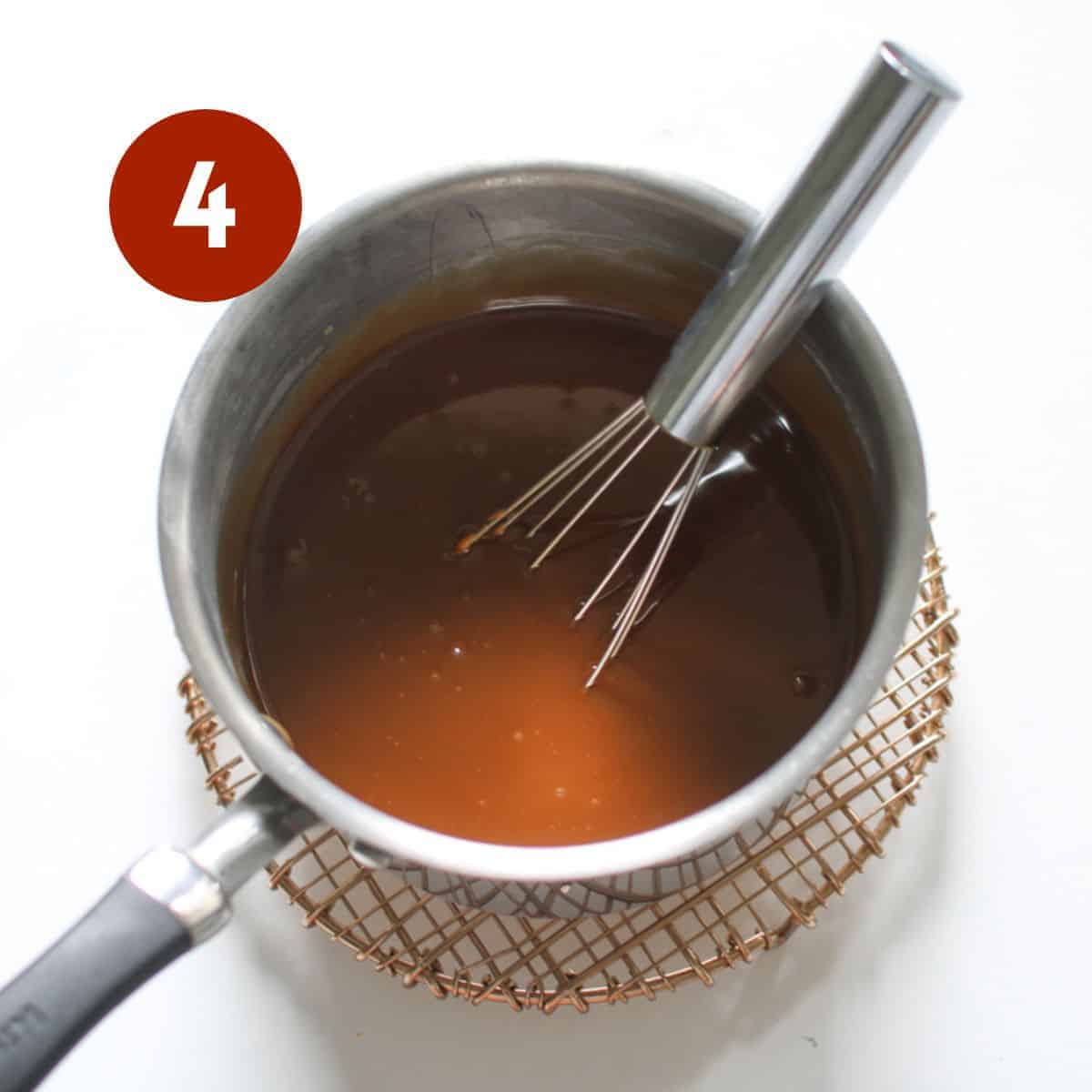 A small sauce pot with a shiny brown cookie butter sauce on a small rose gold trivet on a white background and a metal whisk in the mixture. A white '4' in a dark red circle in the upper left corner.
