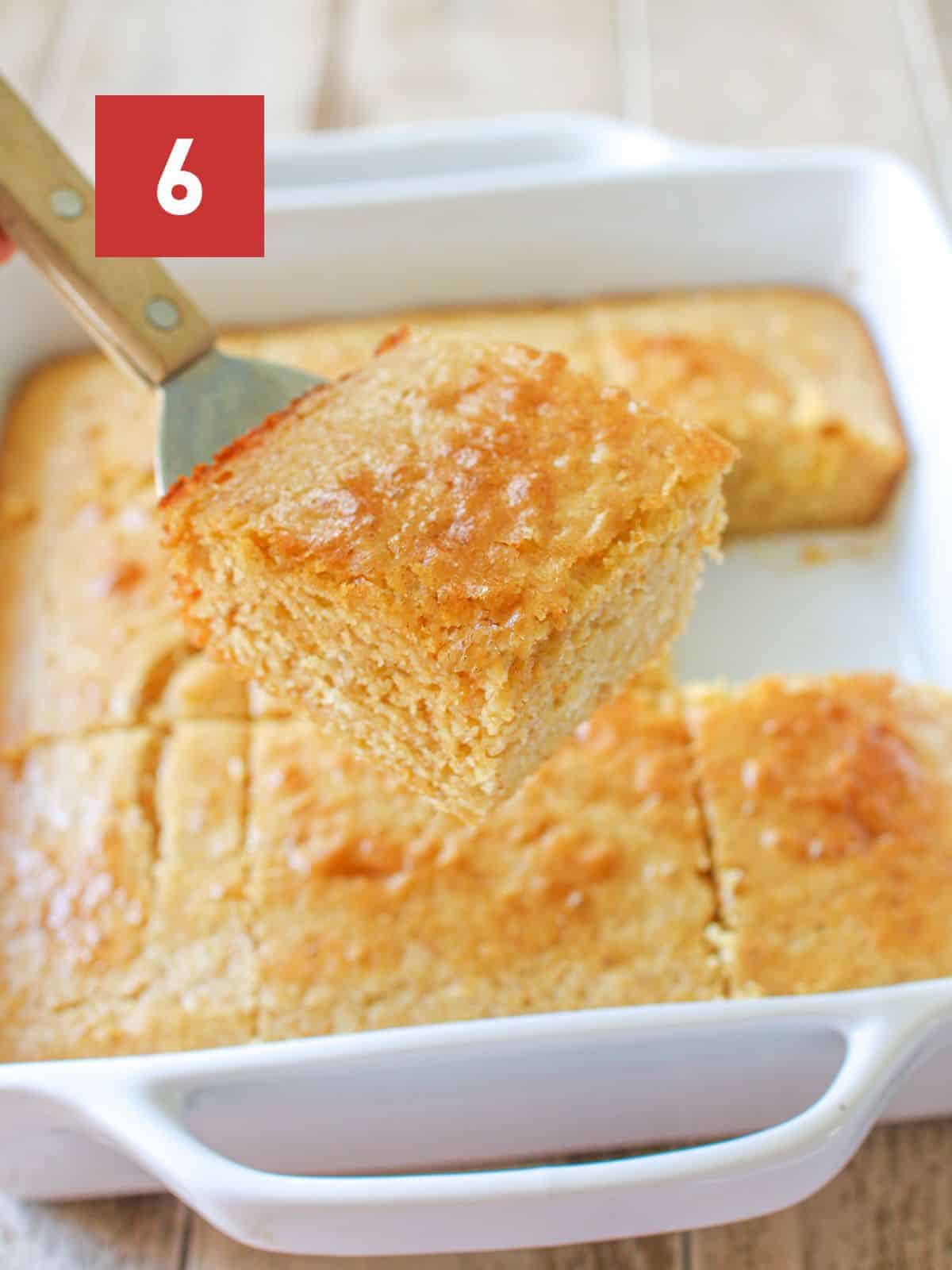 A hand holding a small spatula with a piece of cornbread on it. In the background is a square white baking pan with the rest of the cornbread on a light wood background. A red square box is in the upper left corner with a white '6' in the center.
