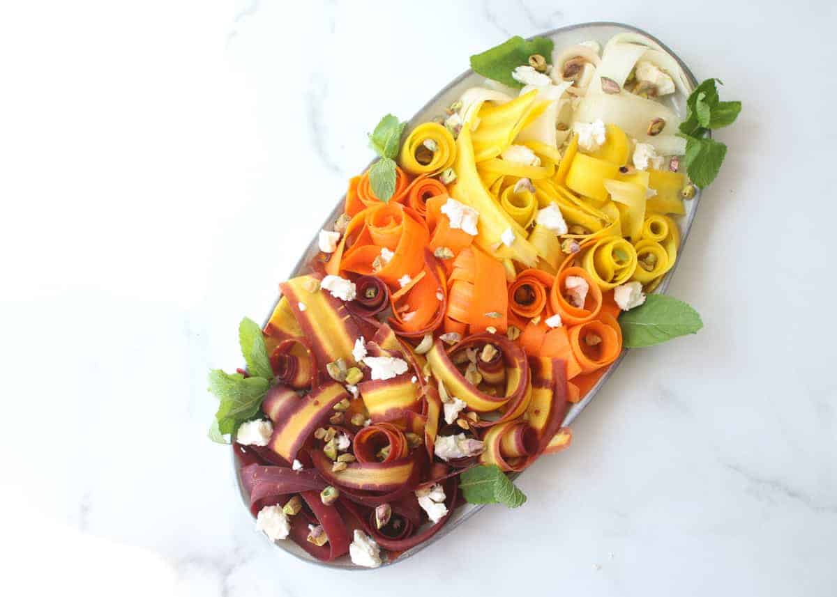 Overhead Rainbow Carrot Ribbon Salad on oval platter with mint on side, topped with feta and pistachios. Carrots in ombree from purple to orange to yellow to white