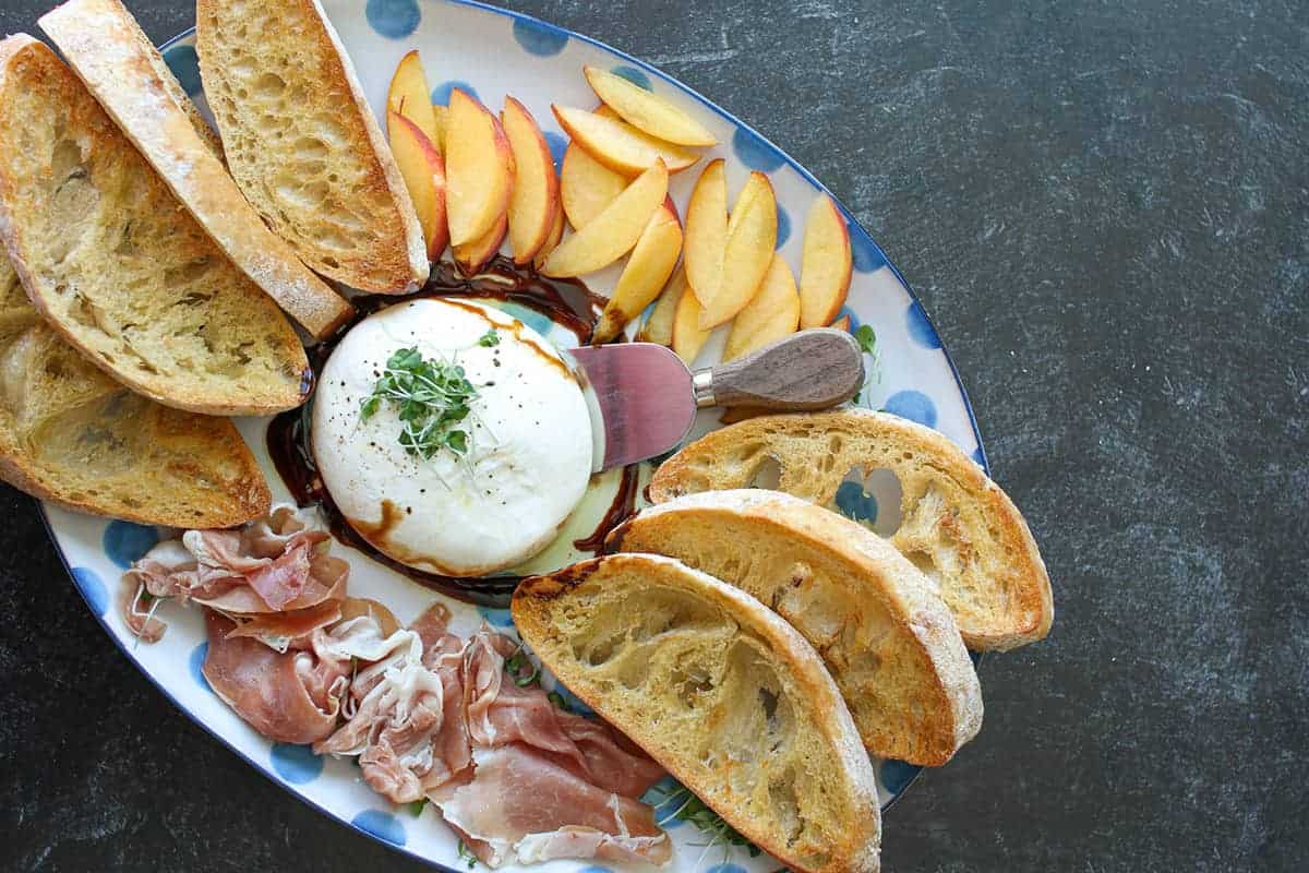 Overhead of a close up of A hand takes a piece of bread off a Burrata, Prosciutto, and Peaches Plate on a black background. There's a small wood cheese knife by the burrata.