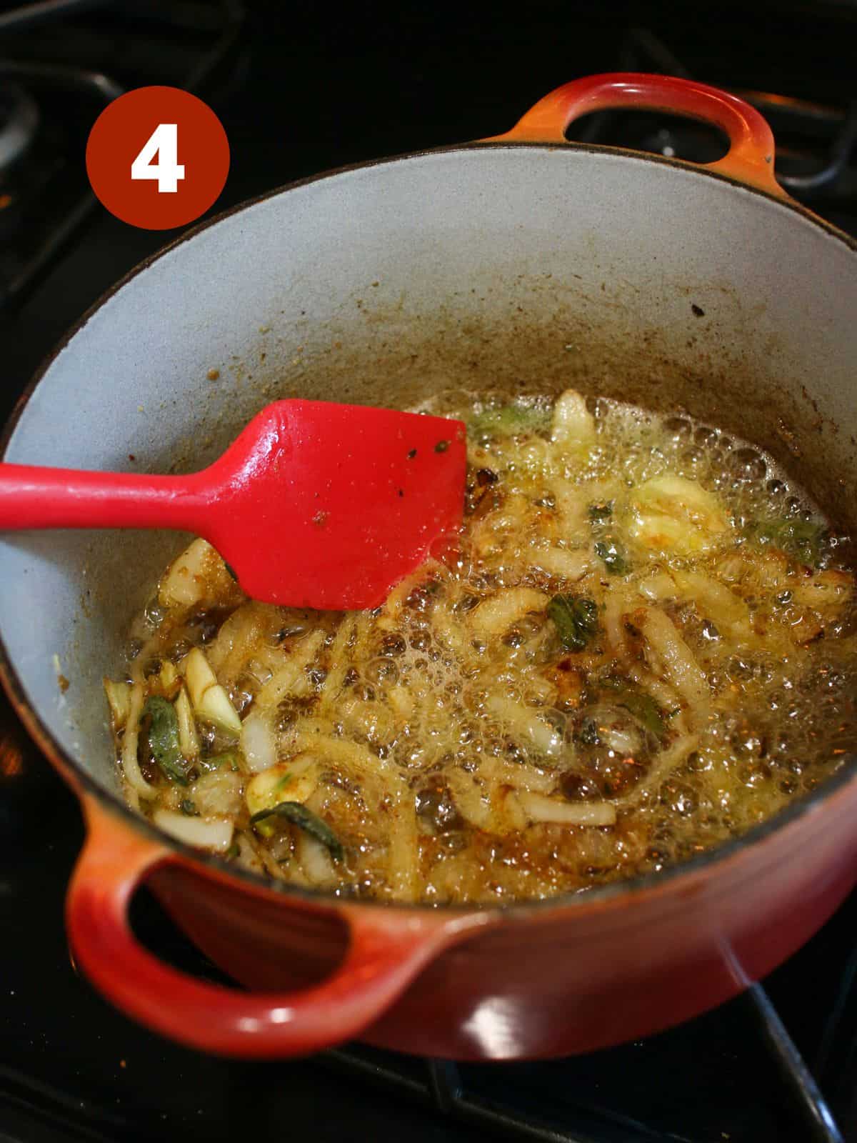 A small red dutch oven on the stove with sliced onions, oil, fresh sage and thyme, crushed garlic, pepper and chili flakes all mixed together with lots of butter bubbling. A red silicone spatula mixing the onions together. In the upper left corner is a '4' in a dark red circle.
