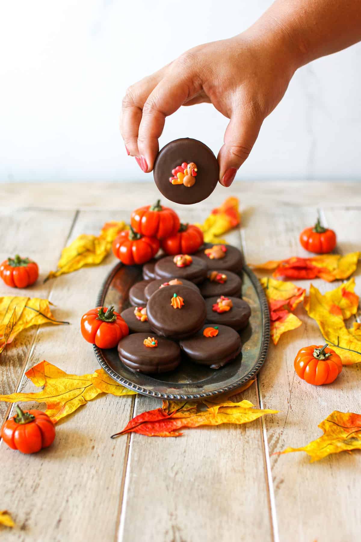 A small antique tray with Thanksgiving Chocolate Covered Oreos stacked on top surrounded by mini pumpkins and yellow/orange leaves on a wood plank surface with white marble in the background. A tan hand picks up an oreo with a decorated turkey on top.