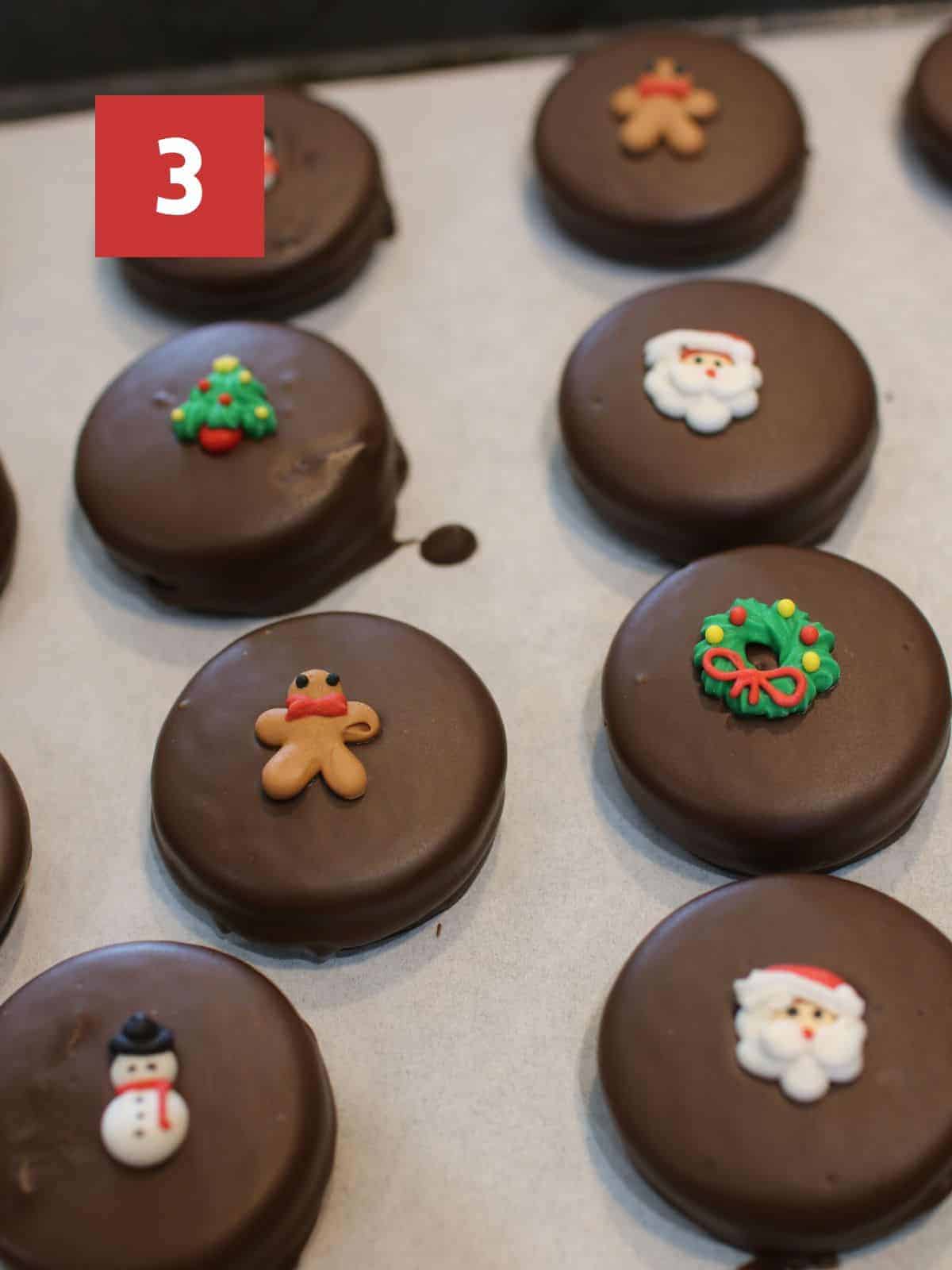 Chocolate covered Oreos with either a Christmas royal icing piece on top on a white piece of parchment paper. In the upper left corner is a dark red square with a white '3' in the center.