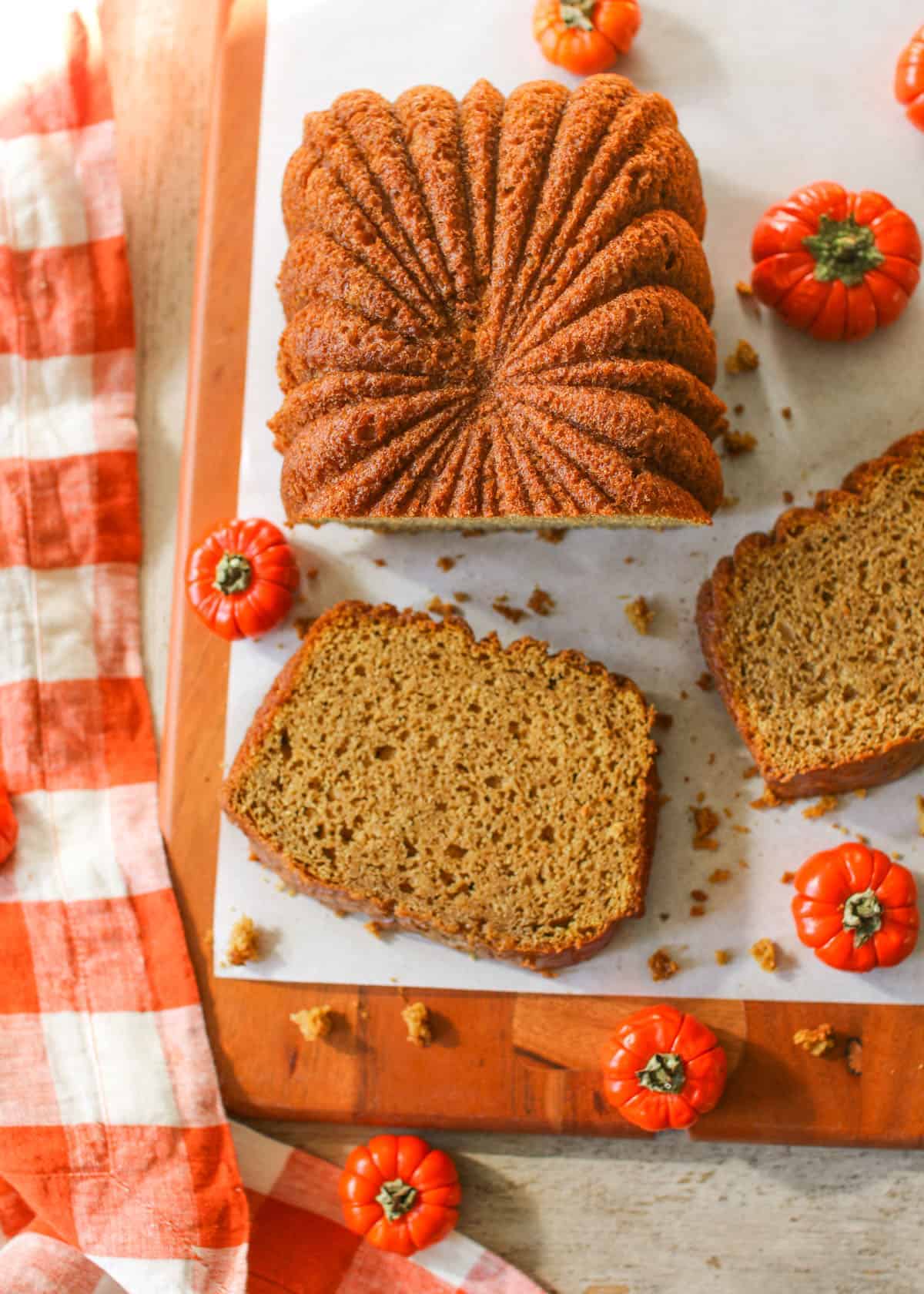 Sliced High Altitude Pumpkin Bread on a wooden board lined with white parchment paper. Surrounded by mini orange pumpkins on a wood background.