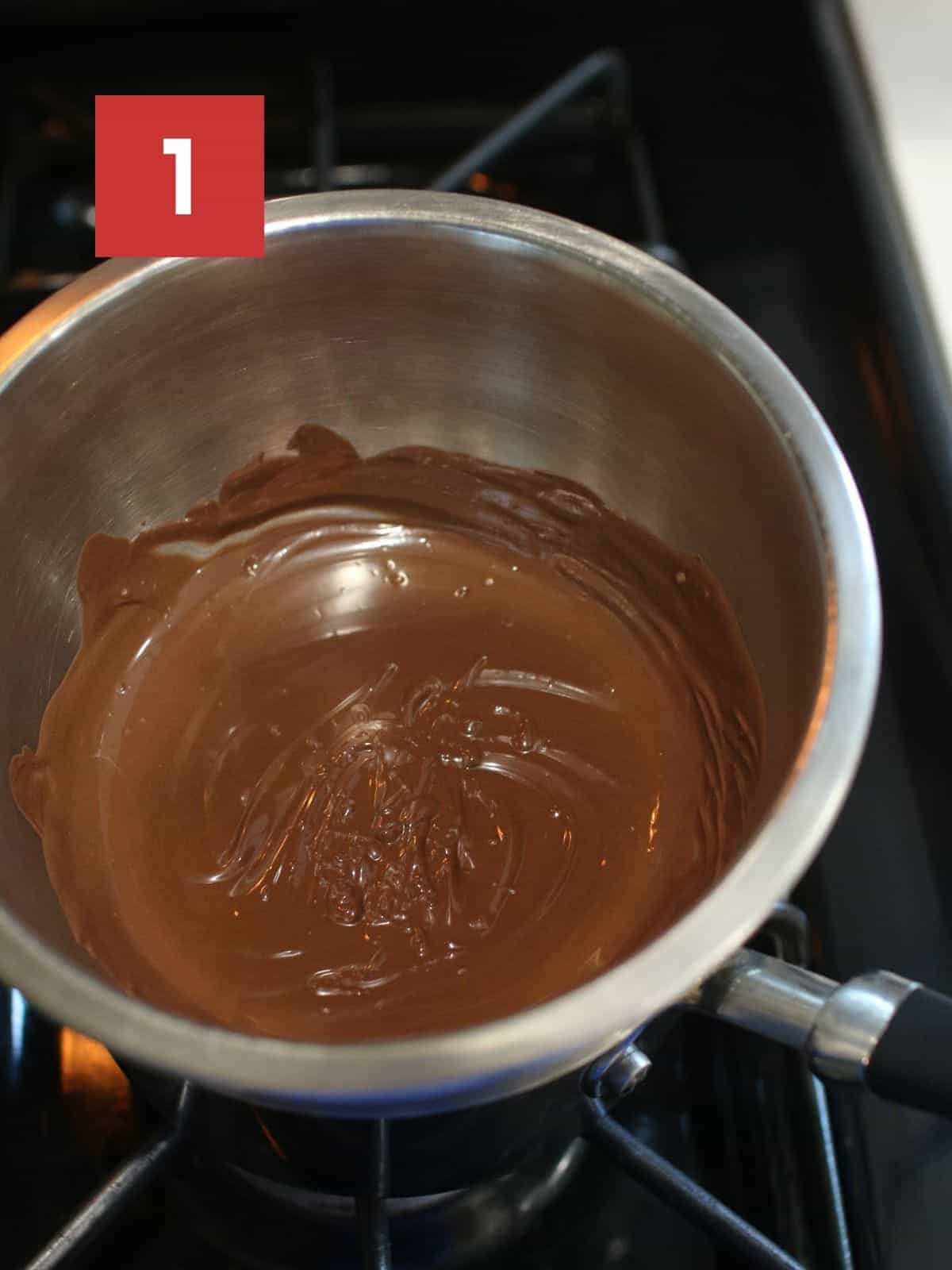 A small metal bowl set up on a double broiler on the stove with melted chocolate. In the upper left corner is a dark red square with a white '1' in the center.