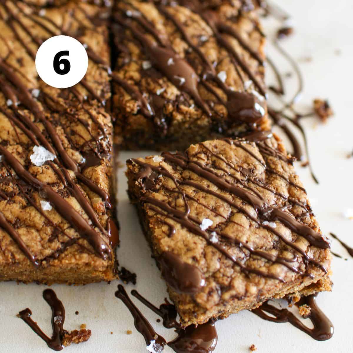 Chocolate Chunk Cookie Butter Bars on a white marble background with one bar slightly offset. The bars have drizzled chocolate and flaky sea salt on top. In the upper left corner is a white circle with a number '6' in black in the center.