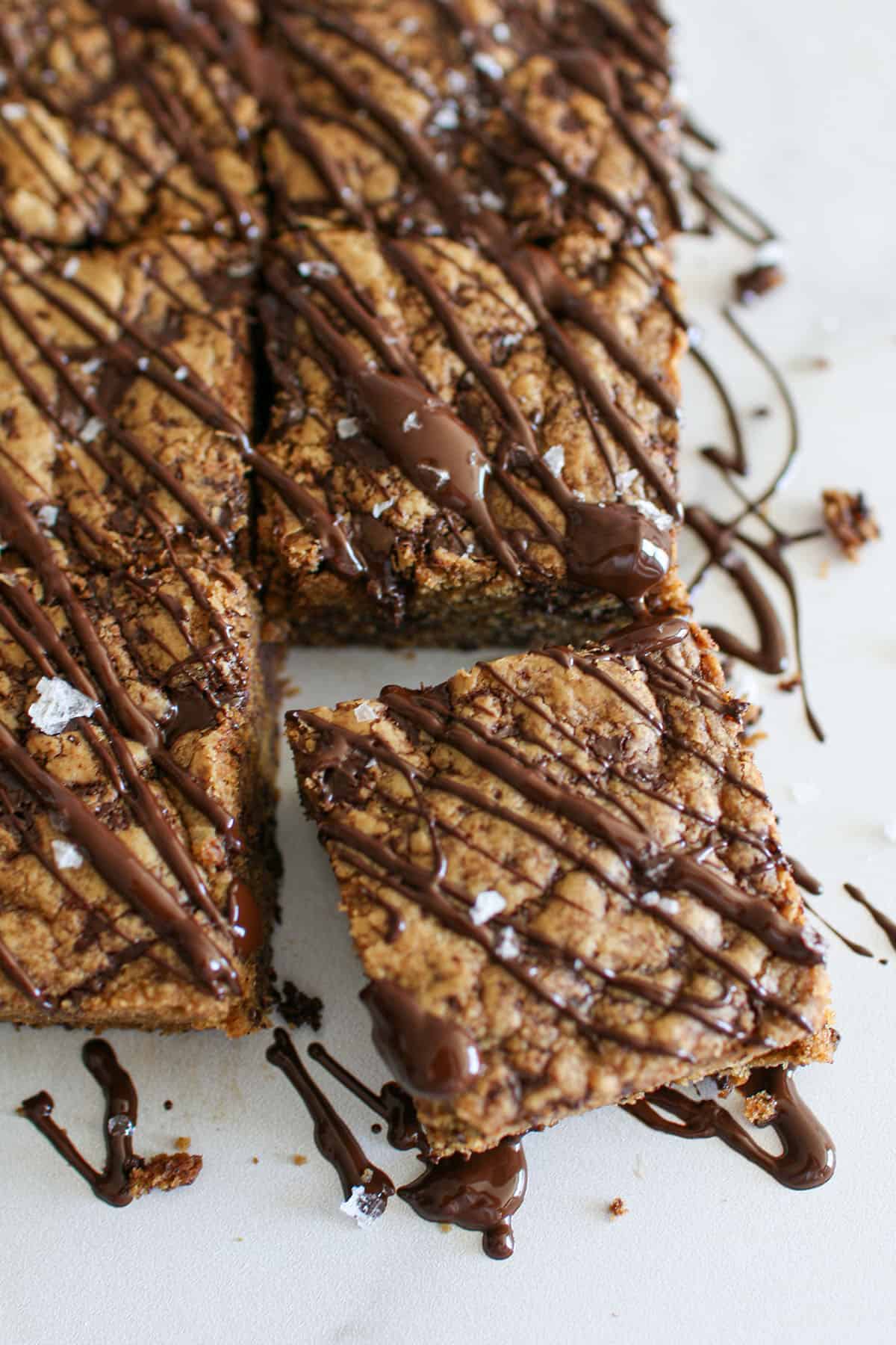 Chocolate Chunk Cookie Butter Bars on a white marble background with one bar slightly offset. The bars have drizzled chocolate and flaky sea salt on top.