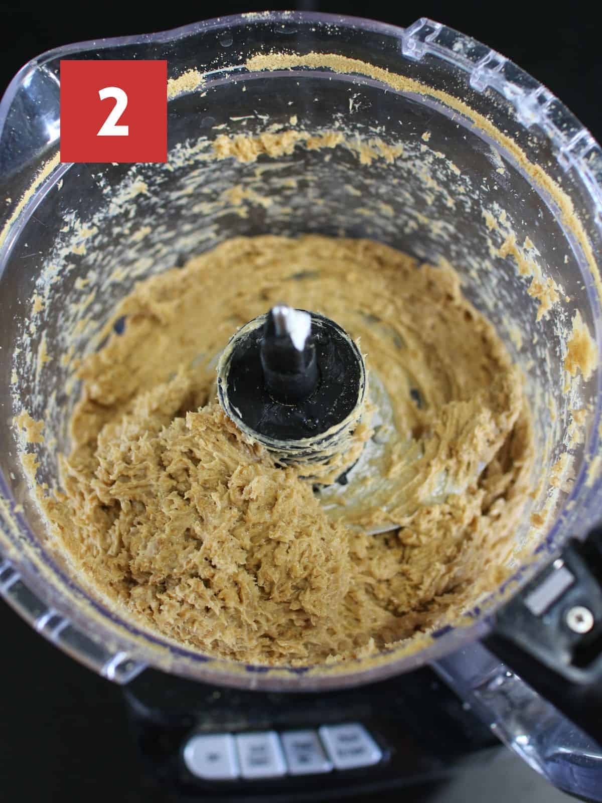 A large food processor with ground ginger snap cookies and cream cheese with top removed on a black table.  In the upper left corner is a dark red square and a white '2' in the center.