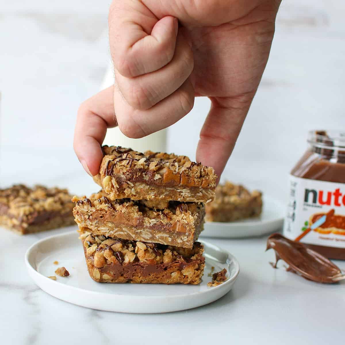 A hand picking up a bar of Nutella Cookie Butter Oatmeal Bar. There's a stack of 3 bars on a small white plate. with milk bottle full of milk and a jar of nutella in the background