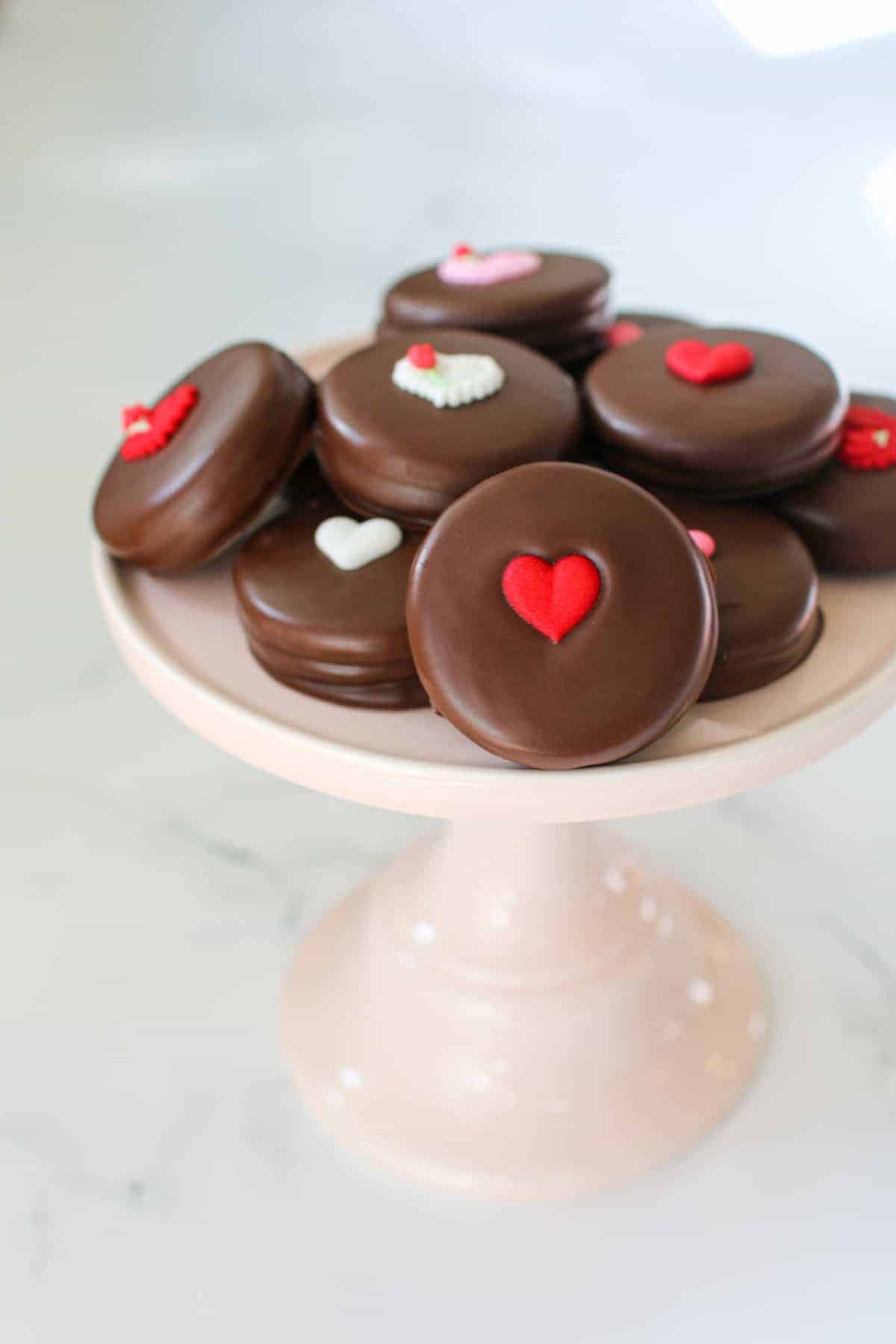 Chocolate covered Oreo cookies with heart decorations on top on a light pink cake stand on a white marble background.