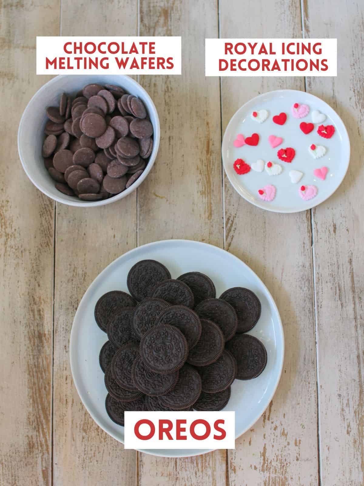A white plate with oreos stacked, a smaller white plate with a variety of heart royal icing pieces and bowl of chocolate melting wafers. Each item is labeled with a white rectangle box with text in all caps in dark red on a light wood plank background.