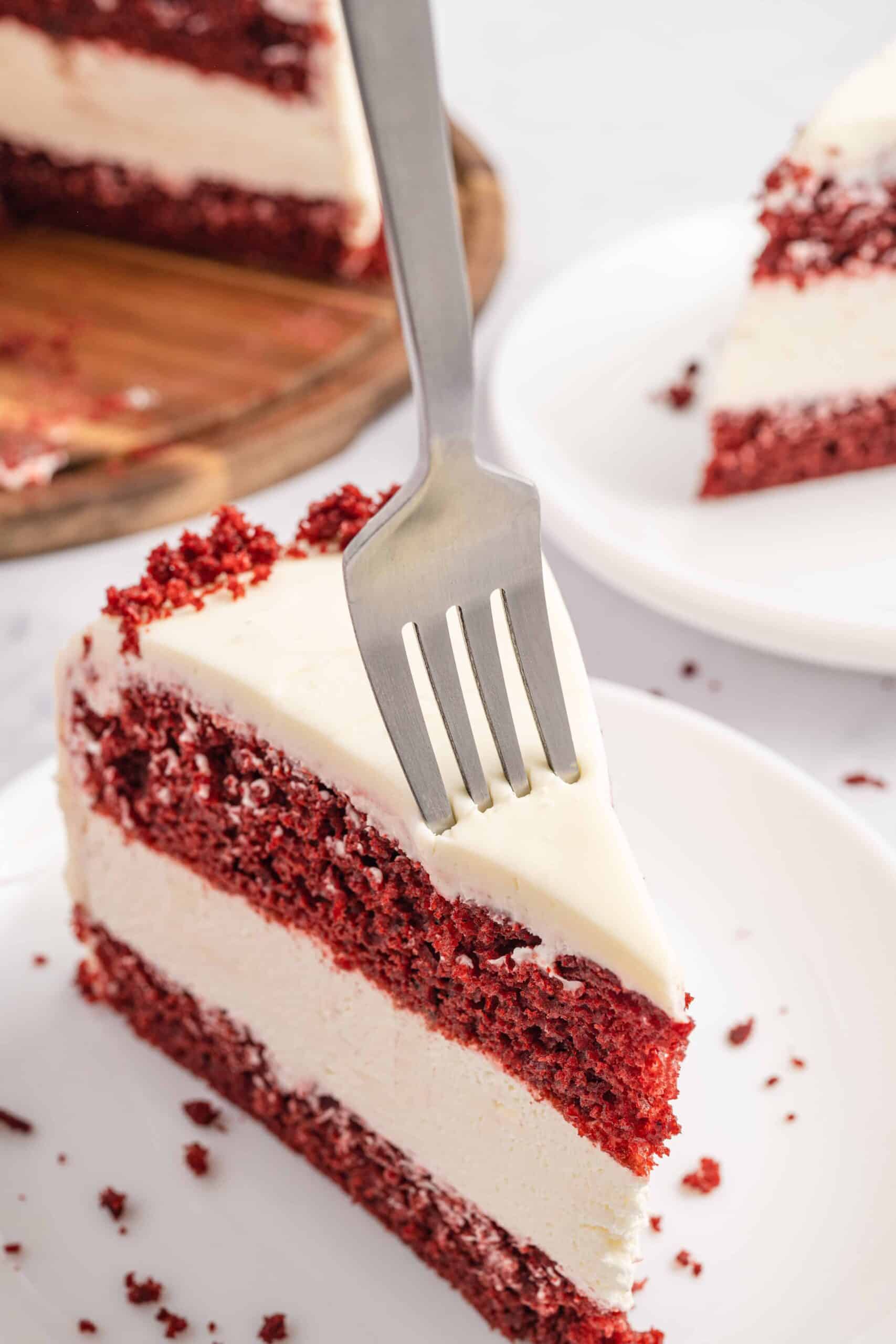 A single slice of Red Velvet Cheesecake Cake on a white plate with the rest of the cake and another slice in the back. In the front slice of cake a silver fork is in in the cake slice.