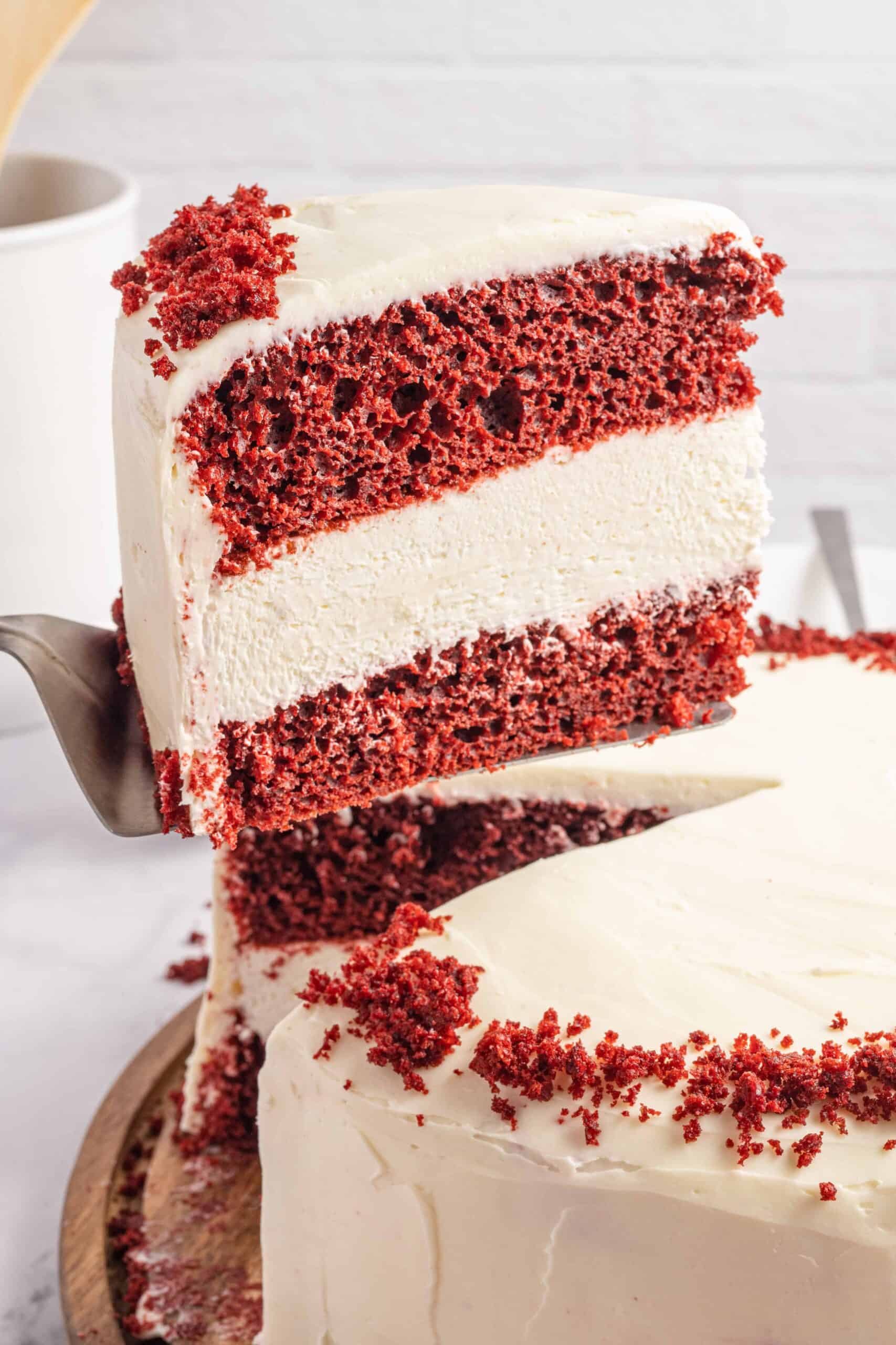 A slice of Red Velvet Cheesecake Cake being recommend from the whole cake on a cake spatula.