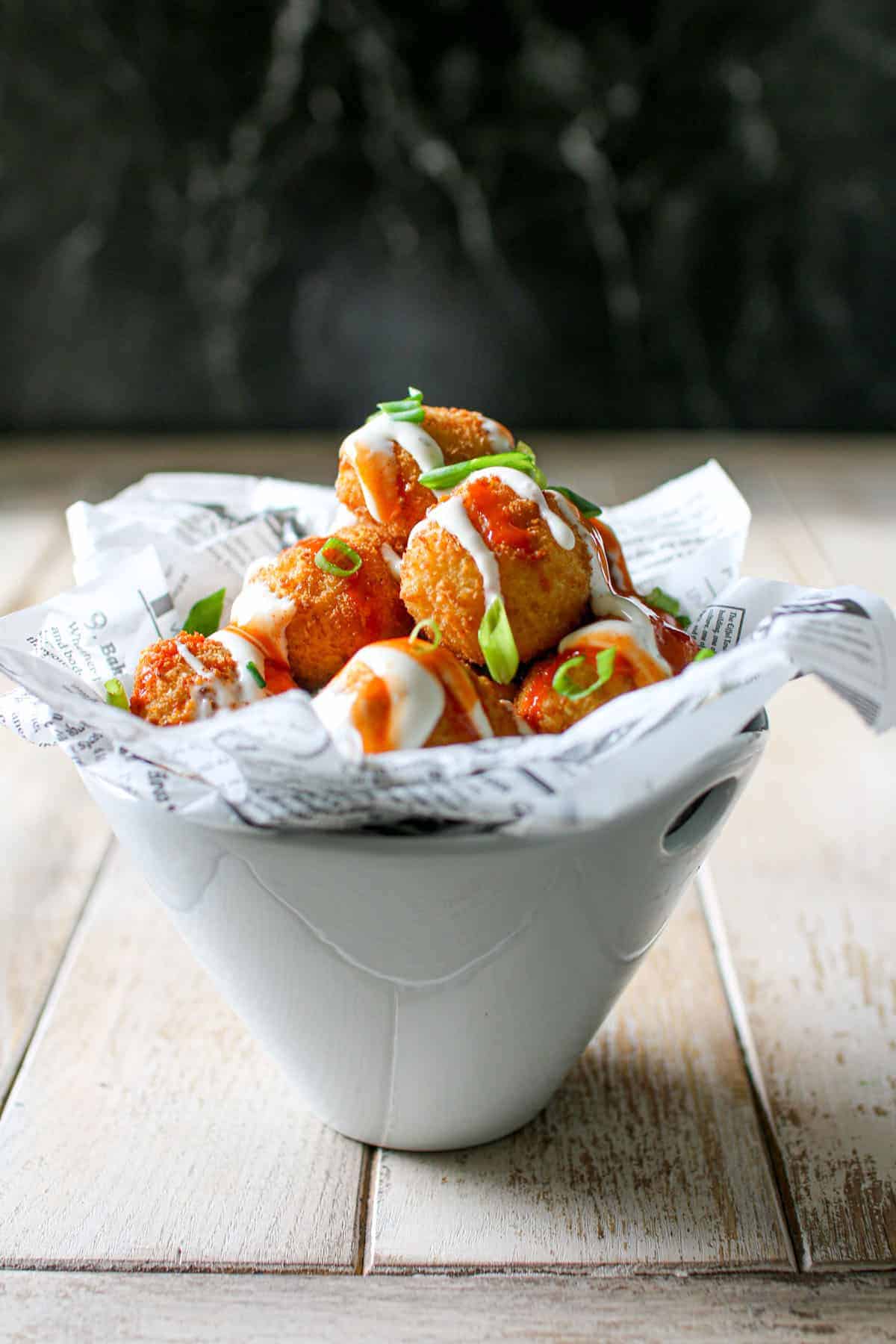Air Fryer Trader Joe's Mac and Cheese Bites stacked in a white bowl and topped with buffalo and ranch dressing with green onion on top. It sits on a wood board with a black marble background.