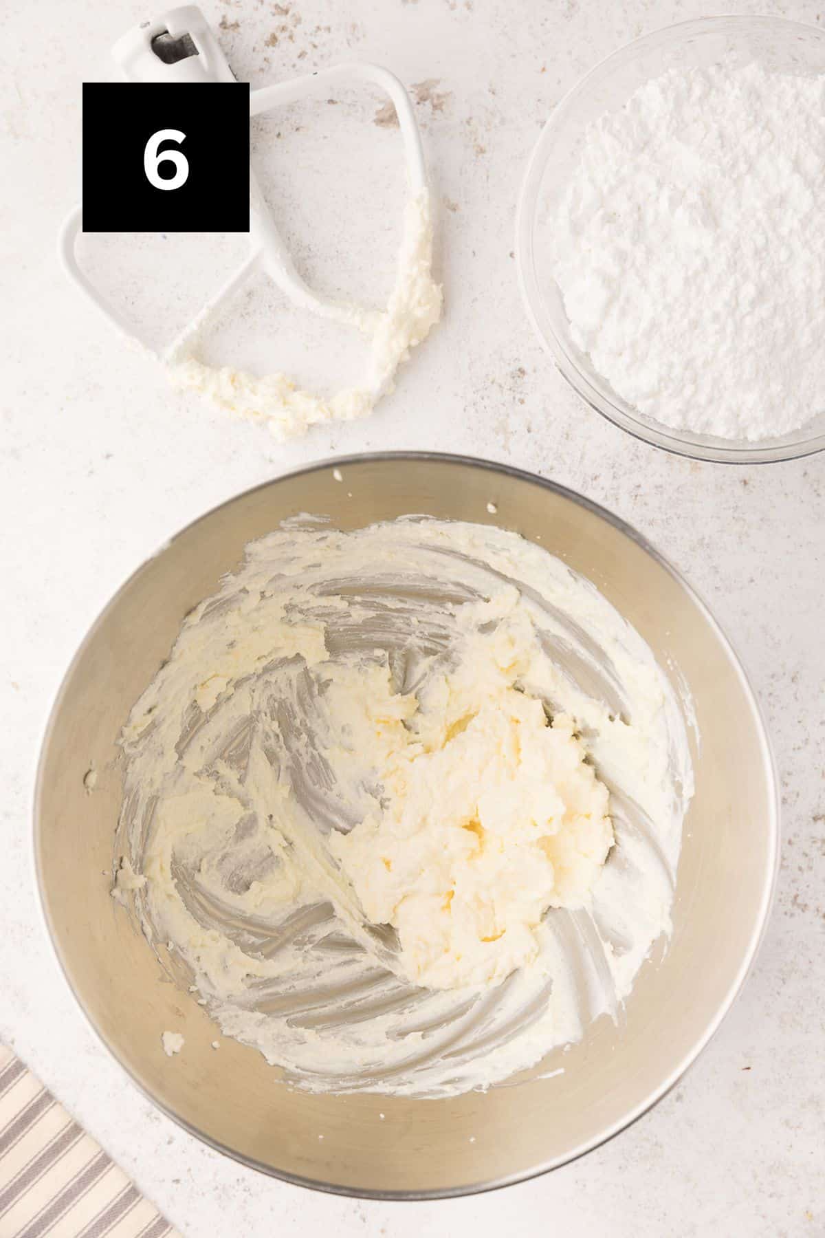 Cheese cheese and butter beaten in a steel mixing bowl with a bowl of powdered sugar off to the side. In the upper left corner is a black square with a white '6'. 