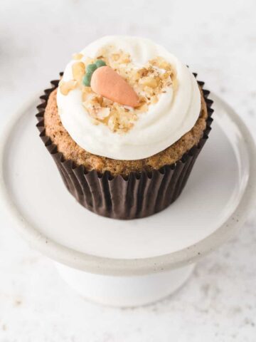 A single Carrot Cake Cupcake on a white mini cake stand. The cupcake has a dark brown cupcake liner and is topped with cream cheese frosting, chopped walnuts and a gum paste carrot.