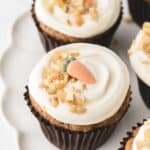 Several Carrot Cake Cupcakes in a dark brown liner on a white scalloped platter. Each cupcake is topped with cream cheese frosting, chopped walnuts and a gum paste carrot.