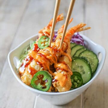 A hand holding chopsticks picking up a piece of shrimp tempura. A white bowl with Frozen Tempura Shrimp in Air Fryer with sriracha mayo, sweet soy sauce, thinly sliced red and green cabbage, cucumber, rice, thin jalapeno and white sesame seeds.