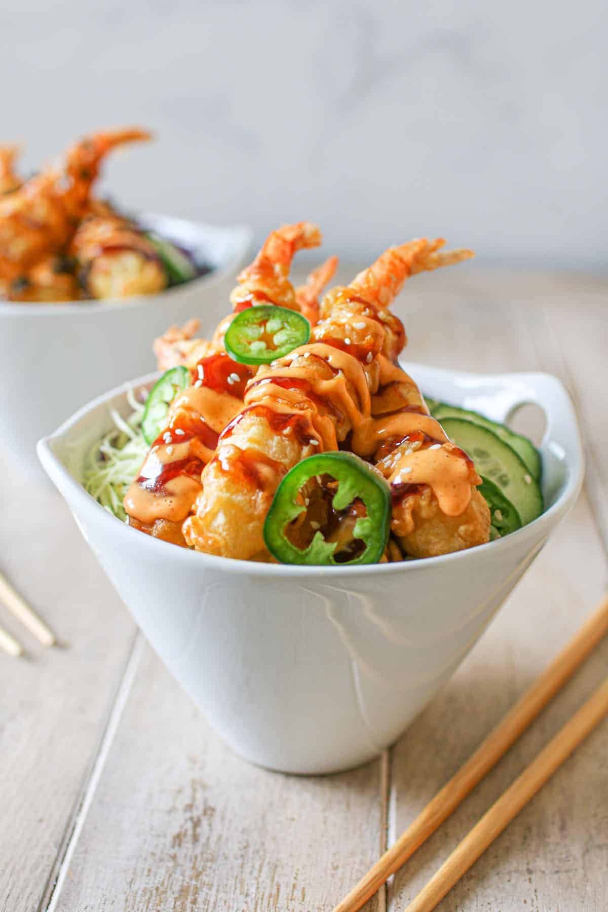  A white bowl with Frozen Tempura Shrimp in Air Fryer with sriracha mayo, sweet soy sauce, thinly sliced red and green cabbage, cucumber, rice, thin jalapeno and white sesame seeds. Another bowl is off in the background with 2 pairs of chopsticks on a wood table and a white marble background.