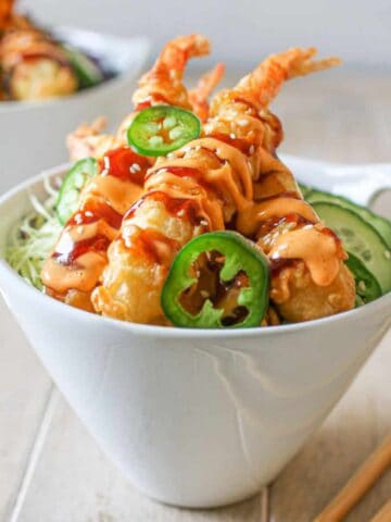 A white bowl with Frozen Tempura Shrimp in Air Fryer with sriracha mayo, sweet soy sauce, thinly sliced red and green cabbage, cucumber, rice, thin jalapeno and white sesame seeds. Another bowl is off in the background with 2 pairs of chopsticks on a wood table and a white marble background.