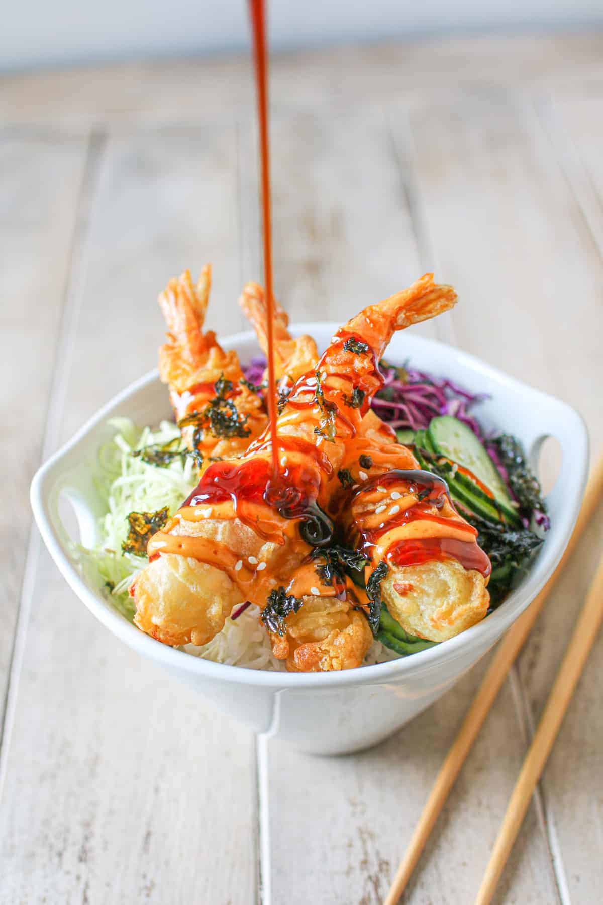 A white bowl with Frozen Tempura Shrimp in Air Fryer with sriracha mayo, sweet soy sauce, thinly sliced red and green cabbage, cucumber, rice and seaweed. Thick sweet sauce is being drizzled on top and a pair of chopsticks on the side.
