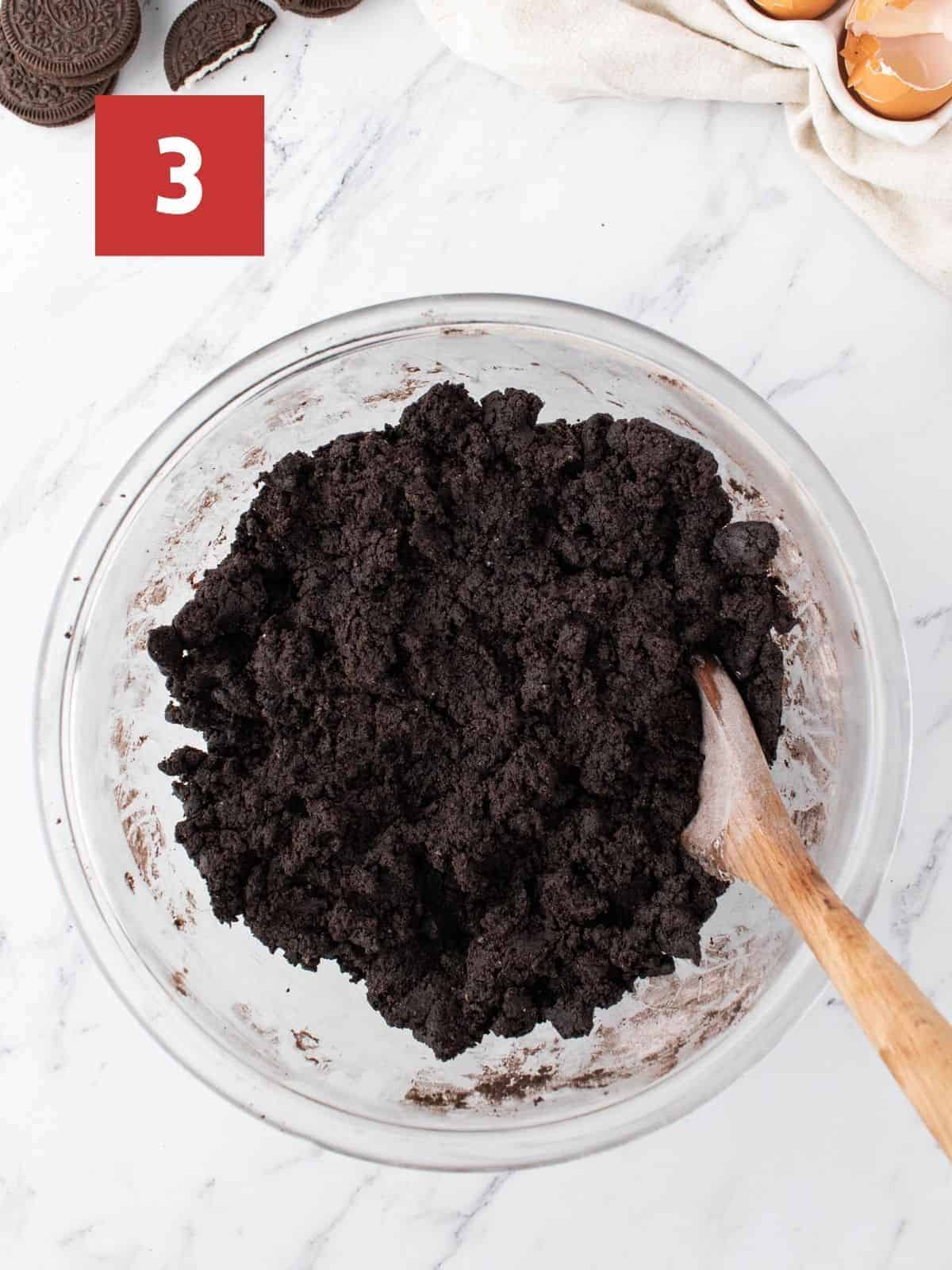 A glass mixing bowl with a dark brown/black cookie dough with a wooden spoon in the mix. The bowl sits on a white marble background with a red square in the upper left corner with a white '3'.