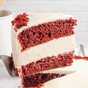 A slice of Red Velvet Cheesecake Cake being recommend from the whole cake on a cake spatula.